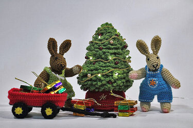 Zwei gehäkelte Amigurumi-Hasen neben einem Tannenbaum mit Lichterkette; roter Häkelwagen mit Geschenkpäckchen.