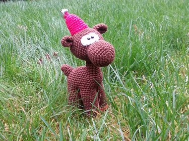 Gehäkeltes braunes Amigurumi-Rentier mit pinker Zipfelmütze und Glöckchen im Gras