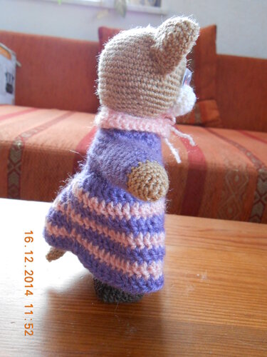 Gehäkelte Amigurumi-Katze, seitliche Ansicht, lila-pink gestreiftes Kleid und rosafarbener Kragen, steht auf einem Holztisch.