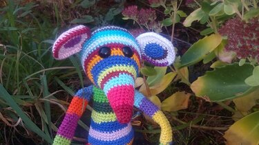 Bunte gestreifte Amigurumi‑Maus mit großen Ohren vor Gartenpflanzen