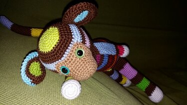 Nahaufnahme eines gehäkelten Amigurumi-Äffchens in leuchtenden Farben auf grünem Stoff, mit großen Augen und gestreiften Gliedmaßen.