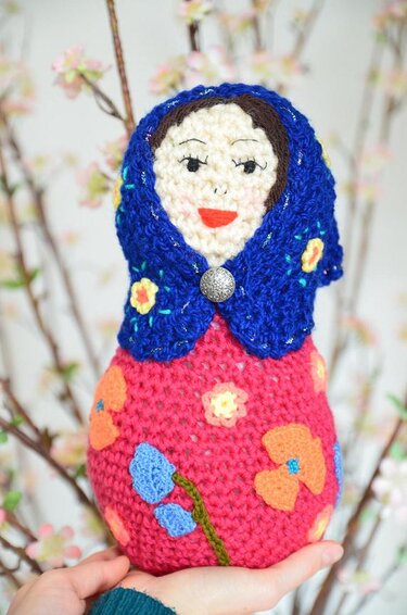 Gehäkelte Amigurumi-Matroschka mit blauem Kopftuch und pinkem Körper mit Blumenapplikationen