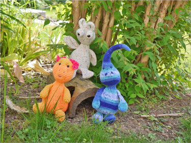 Drei gehäkelte Amigurumi: orangefarbenes Wesen mit rosa Schleifen, gesprenkelter Hase und blau gestreiftes Fantasiewesen vor einem Baumstamm im Gras
