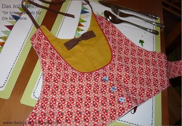 Babylätzchen mit rot-rosa geometrischem Muster, senffarbenem Futter, Paspel, drei Knöpfen und Bindebändern, dekoriert mit einer kleinen Fliege.