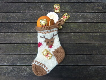 Gestrickter Weihnachtsstrumpf in Weiß und Braun mit Rentier-Motiv, gefüllt mit Orange und Schokolade auf Holzoberfläche.