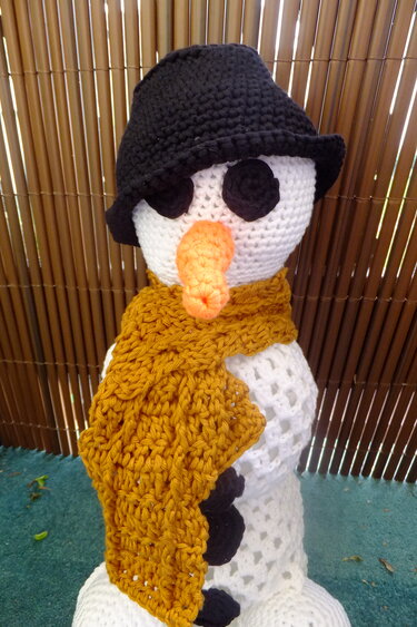 Gehäkelter Schneemann in Weiß mit schwarzem Hut, senfgelbem Schal, orangefarbener Karottennase und schwarzen Knöpfen vor einer Bambuswand.