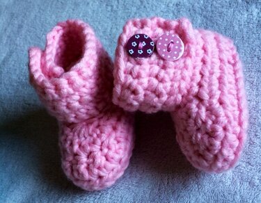 Zwei rosa gehäkelte Babyschuhe mit Umschlag und zwei Zierknöpfen auf hellem Stoff