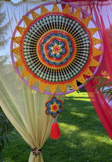 Bunter gehäkelter Traumfänger mit sonnenförmigem Mandala-Muster in einem lila Ring, mit angehängter Häkelblüte und roter Quaste