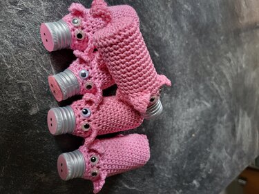 Mehrere rosa gehäkelte Überzüge mit aufgesetzten Augen und Knopf‑Nasen auf Schraubverschlüssen