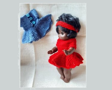 Dunkelhäutige Puppe in rotem Häkelkleid und Stirnband, daneben blaue Häkeljacke mit Knöpfen