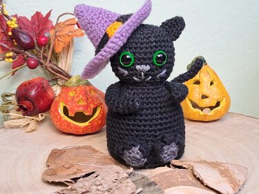 Schwarze Amigurumi-Katze mit grünen Augen und lila Hexenhut vor Halloween-Deko-Kürbissen