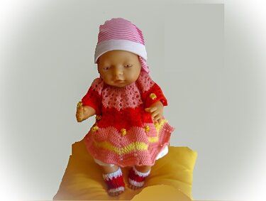 Babypuppe auf gelbem Kissen trägt gehäkeltes rosa-rotes Kleid mit gelben Wellenstreifen, gestreifte Mütze und kleine Söckchen