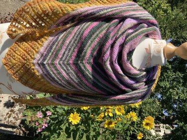 Mehrfarbiges gestricktes Dreieckstuch mit senfgelbem Lochmuster und bunten Streifen, drapiert auf einer Büste im Garten.