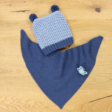 Hellblaue gestrickte Baby-Mütze mit zwei Bommeln neben dunkelblauem gestricktem Dreieckstuch auf Holzuntergrund