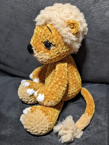 gelb‑beiger gehäkelter Amigurumi‑Teddy mit plüschiger Mähne und Schwanzquaste, seitliche Ansicht, sitzend