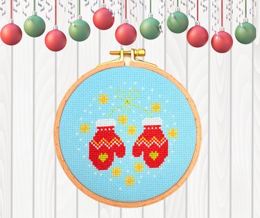 Rundes Kreuzstichbild mit roten Fäustlingen auf hellblauem Stoff im Holzstickrahmen vor weißer Holzoberfläche mit Weihnachtskugeln