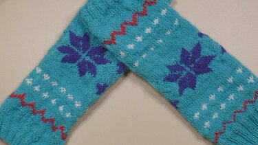 Zwei gestrickte türkisfarbene Armstulpen mit lila Schneeflocken und roten Zickzackmustern