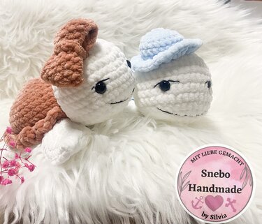 Zwei kompakte weiße Amigurumi-Wasserfiguren aus Plüschgarn, eines mit braunen Details und eines mit blauem Häubchen, auf weißem Fell