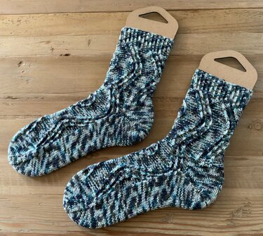 Paar gestrickter Socken mit Zopfmuster und meliertem Blau‑Türkis auf Holzuntergrund