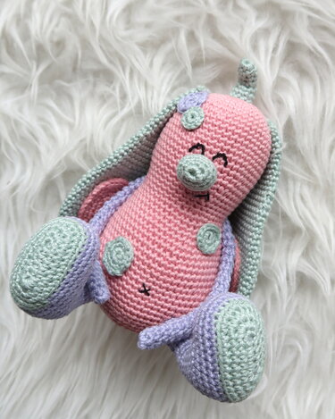 Gehäkeltes Amigurumi‑Häschen in Rosa mit mintgrünen Ohren und fliederfarbenen Sohlen auf weißem, flauschigem Untergrund