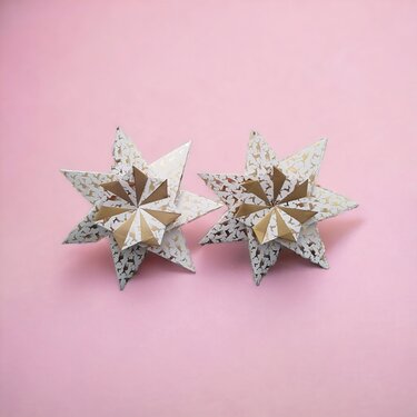 Zwei weiße Papiersterne mit goldenen Rentiermustern auf rosa Hintergrund