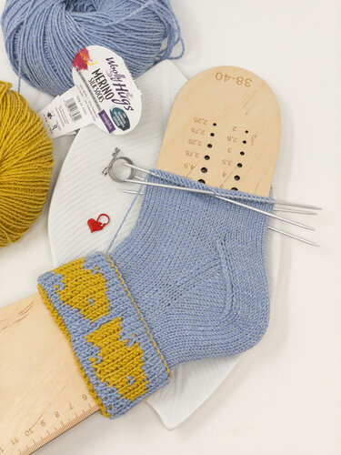 Hellblaue Socke mit gelbem Fischmuster im Bündchen auf Holz-Sockenspannform mit Nadelspiel und Wollknäueln