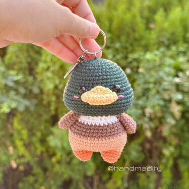 Mini crochet duck keyring with green head, yellow beak, brown body and orange feet held by a hand