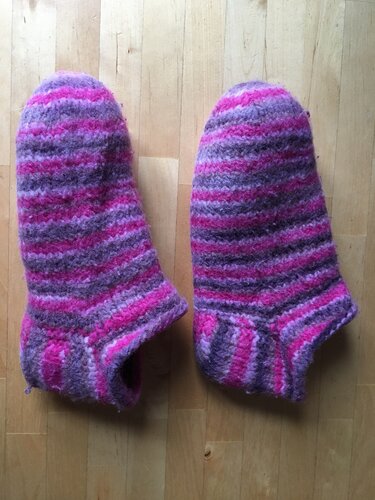 Zwei gestrickte Hausschuhe mit horizontalen Streifen in Pink, Lila und Grau auf Holzboden.
