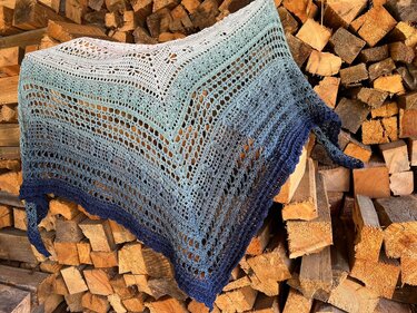 Gehäkeltes Dreieckstuch in Blau- und Türkistönen mit Lochmuster, auf einem Holzstapel drapiert.