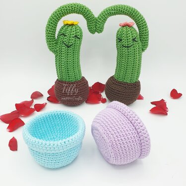 Two green crocheted amigurumi cacti with smiling faces and small flowers form a heart with their arms, with pastel crocheted pots and red flower petals.