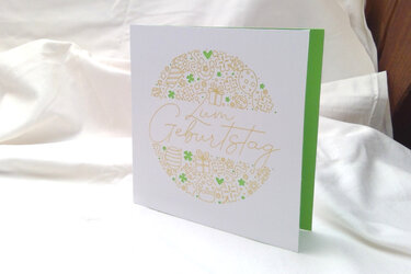 Weiße, quadratische Geburtstagskarte mit goldfarbenem Kreismotiv und grün akzentuierten Glückssymbolen sowie handgeschriebener Aufschrift „Zum Geburtstag“