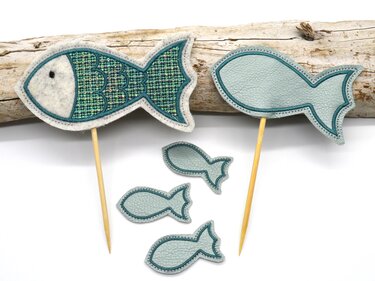 Zwei bestickte Fischfiguren auf Holzstäben und drei kleine Fischapplikationen in Türkis- und Hellblautönen vor einem Stück Treibholz auf weißem Hintergrund.