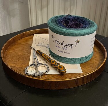 Rundes Holztablett mit Garnknäuel mit Farbverlauf, Holz‑Häkelnadel, verzierter Stickschere und Musterdiagramm