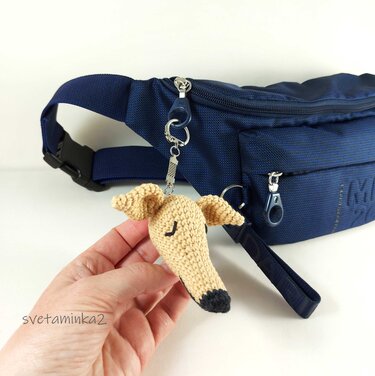 Hand holding a tan crocheted amigurumi dog head keychain clipped to a navy blue waist bag.