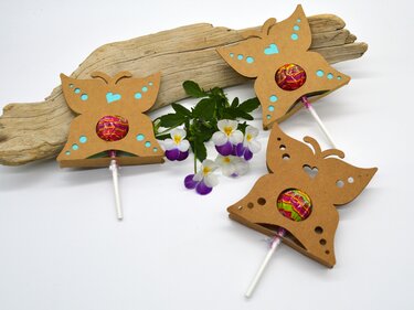 Drei schmetterlingsförmige Lutscherhalter aus braunem Papier, mit bunten Lutschern, neben einem Stück Treibholz und Blüten.
