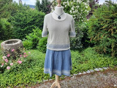 Schneiderbüste trägt beigefarbenen, fein gestrickten Damenpullover mit rundem Ausschnitt, halblangen Ärmeln und hellblauen Kontraststreifen; kombiniert mit blauem Rock vor Gartenkulisse