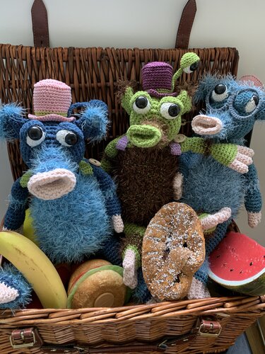 Drei gehäkelte Amigurumi-Monster aus Flauschgarn und Glattgarn sitzen in einem Weidenkorb mit Lebensmitteln.