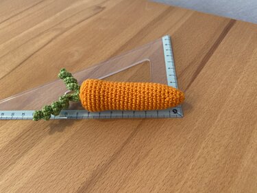 Gehäkelte orange-grüne Karotte neben Lineal auf Holztisch.
