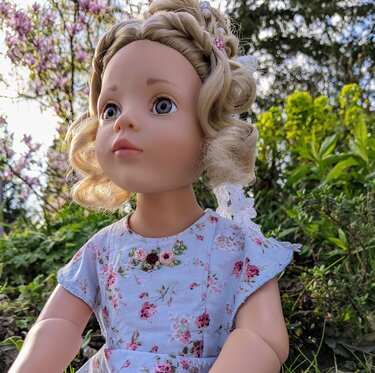 Blonde Puppe mit Locken trägt ein hellblaues Puppenkleid mit Blumenmuster im Garten