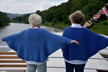 Zwei Frauen in blauen Strickpullover mit weiten Ärmeln auf einem Boot, Blick auf einen Fluss und Bäume.