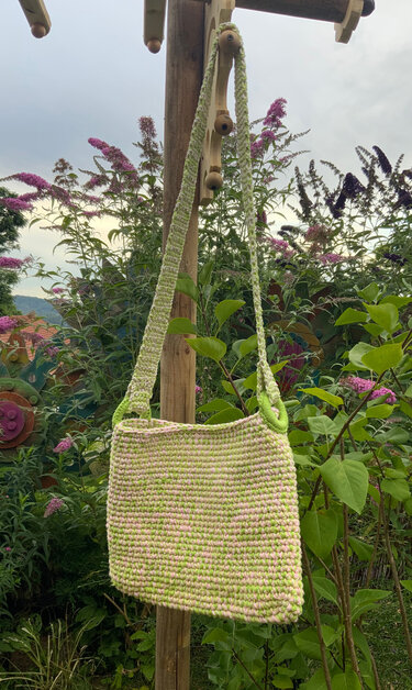 Gehäkelte Umhängetasche in Pastellgrün und Rosa vor blühenden Sträuchern