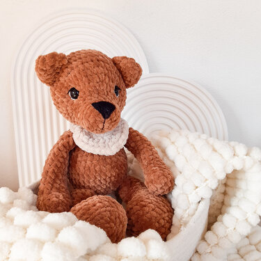 Brauner gehäkelter Amigurumi-Teddybär mit weißem Häkelkragen und schwarzer Nase in heller, kuscheliger Umgebung