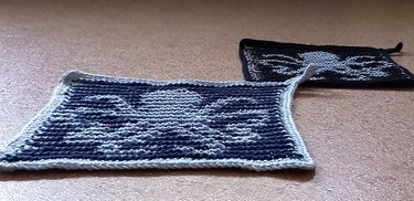 Zwei rechteckige, gestrickte Topflappen mit Totenkopfmuster in invertierten Farbkombinationen und Aufhängeschlaufen.
