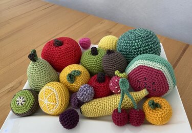 Gehäkeltes Spielobst auf einem weißen Teller: Banane, Kirschen, Kiwi, Zitrusfrüchte und Melone in bunten Garnfarben.