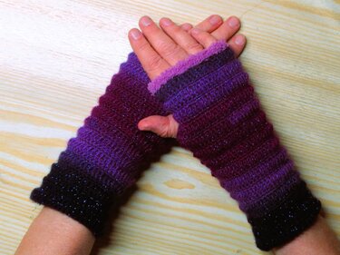 Hände mit gehäkelten fingerlosen Armstulpen in violettem Farbverlauf und Daumenöffnung auf hellem Holzuntergrund