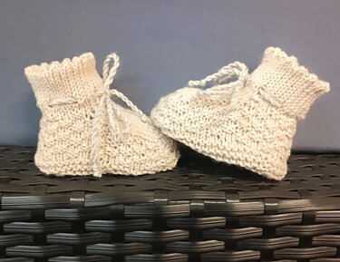 Zwei handgestrickte hellbeige Babyschuhe mit Bindebändern auf dunklem Untergrund