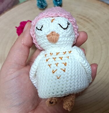 Kleine gehäkelte weiße Amigurumi-Eule mit geschlossenen Augen, gesticktem Brustmuster und rosa Mütze, in einer Hand gehalten.