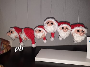 Fünf kleine gehäkelte Weihnachtsmann-Köpfe mit roten Mützen und weißen Bärten an Fäden hängend.