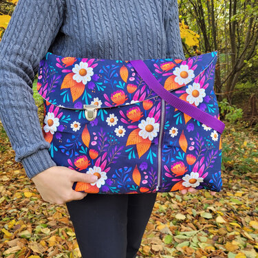 Person hält dunkelblaue Stoff-Umhängetasche mit buntem Blumenmuster, Metallschloss und lila Trageriemen vor herbstlichem Laub.