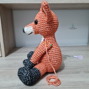 Side profile of orange crochet fox amigurumi with pins holding arm and loose yarn tail